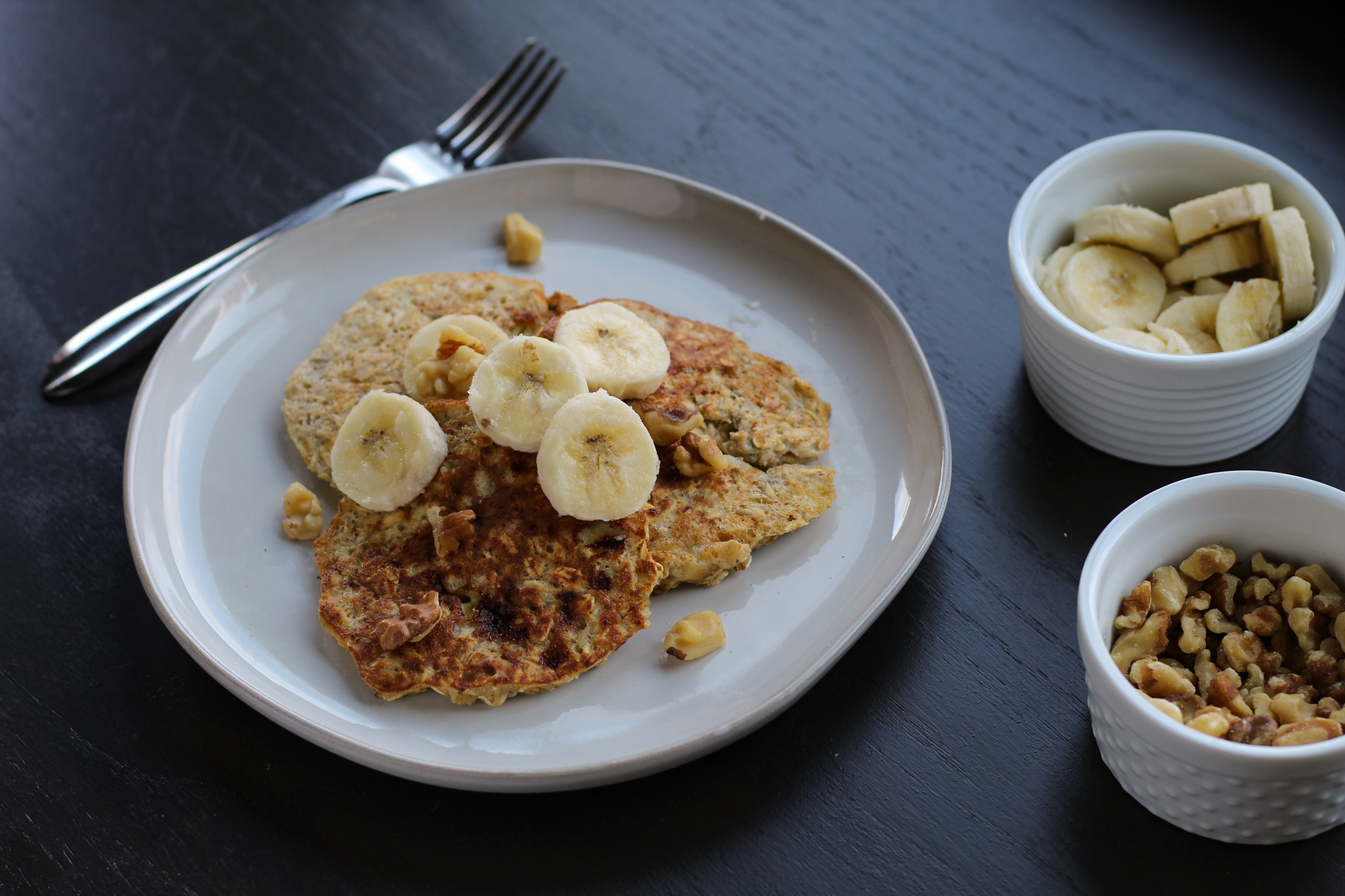 A plate of banana oatmeal pancakes topped with bananas and walnuts next to a fork and bowls of bananas and walnuts.